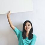 Real Estate Mastery - Cheerful Asian woman sitting cross legged on floor against white wall in empty apartment and showing white blank banner