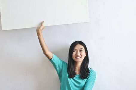 Real Estate Mastery - Cheerful Asian woman sitting cross legged on floor against white wall in empty apartment and showing white blank banner