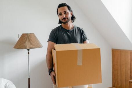 Property Success - Cheerful young bearded male in casual wear carrying cardboard container in sunny bedroom while moving into new apartment
